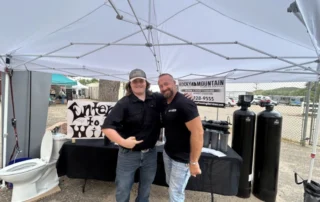 Two men stand smiling under a white canopy at an outdoor event, with water filtration equipment and a toilet on display behind them.