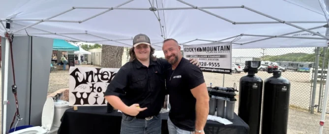 Two men stand smiling under a white canopy at an outdoor event, with water filtration equipment and a toilet on display behind them.