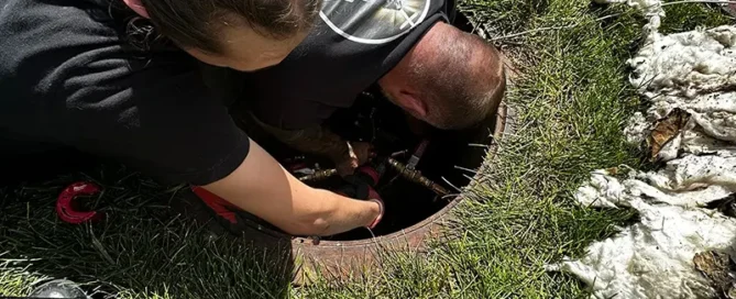 Two people are working together with their arms inside a manhole on a grassy area, surrounded by tools and equipment, demonstrating hands-on Summer Plumbing Tips in action.