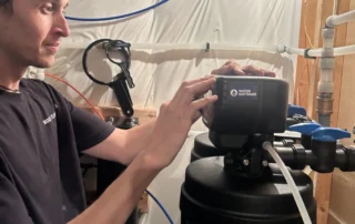 A person adjusts the control panel on a water softener system in a basement or utility room with pipes and tubing visible.