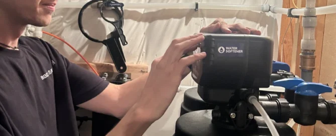 A person adjusts the control panel on a water softener system in a basement or utility room with pipes and tubing visible.