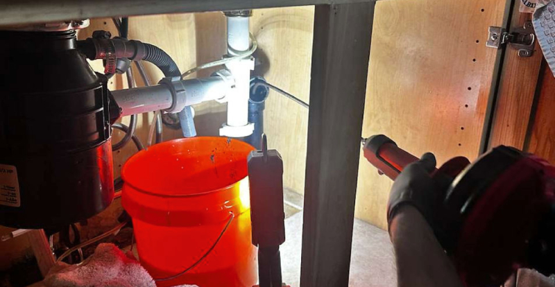 Sink Drain Cleanout Snake A person performs plumbing work, applying sealant to pipes under a kitchen sink, with a red bucket placed below and various plumbing fixtures visible.