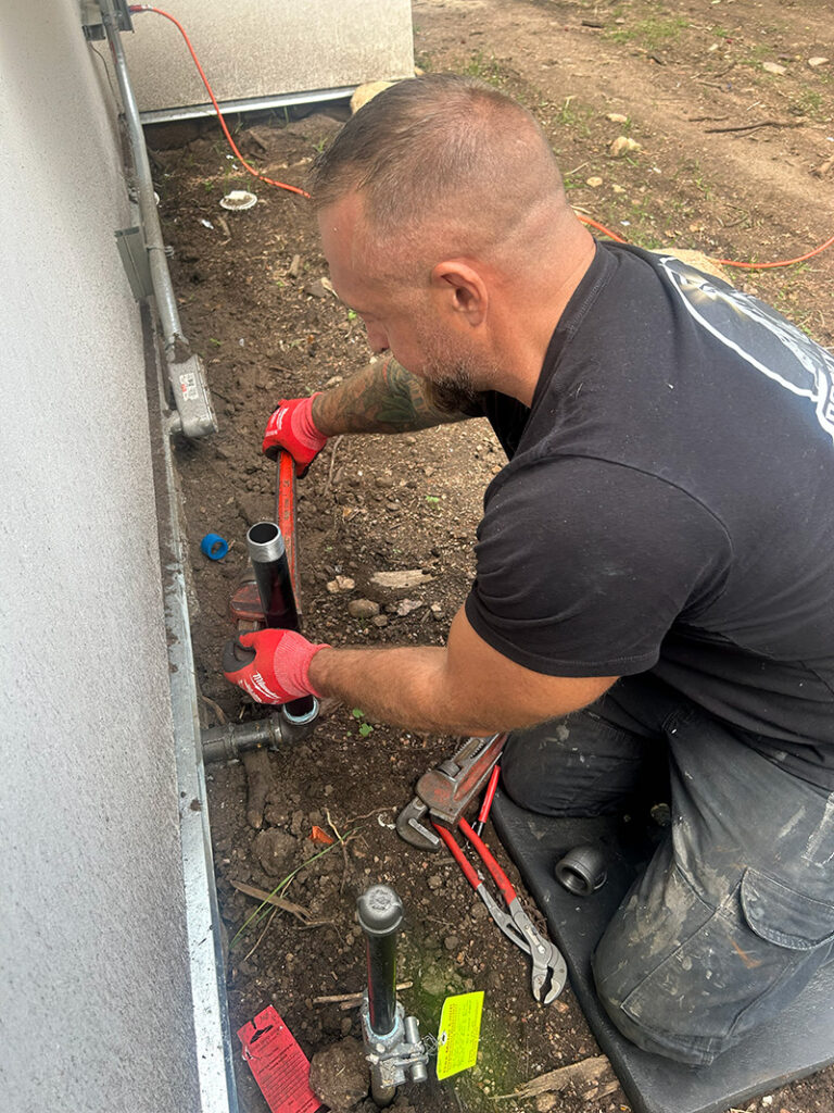 Natural Gas Pipe Install 800 A man kneels on the ground using a red pipe wrench to tighten outdoor plumbing pipes next to a building; various plumbing tools and dirt are scattered around him.