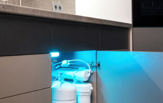A plumbing water filtration system with multiple tanks and pipes is installed under a kitchen sink, illuminated by a blue light.