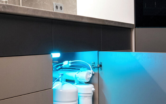 A plumbing water filtration system with multiple tanks and pipes is installed under a kitchen sink, illuminated by a blue light.