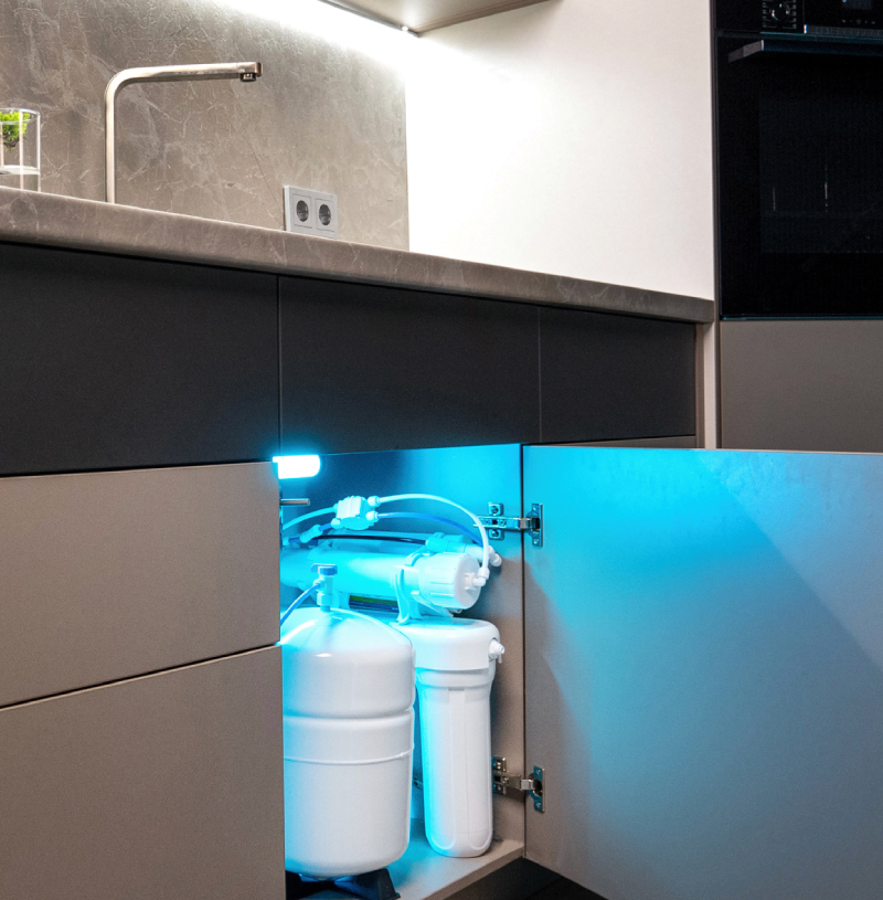 Pure Drinking Water—Right From Your Kitchen Tap A plumbing water filtration system with multiple tanks and pipes is installed under a kitchen sink, illuminated by a blue light.
