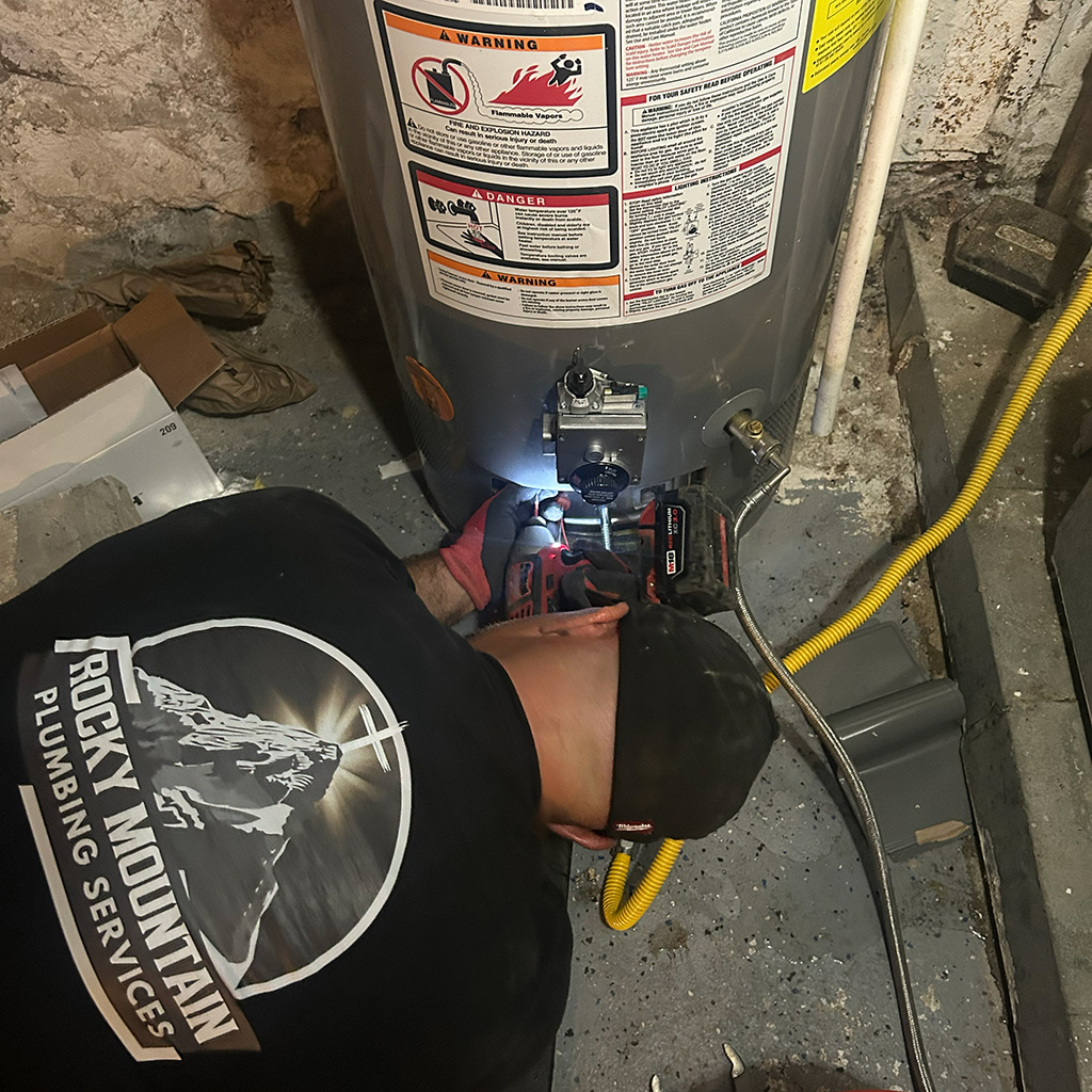A plumber in a "Rocky Mountain Plumbing Services" shirt uses a power tool to handle plumbing repairs at the base of a water heater in a basement.