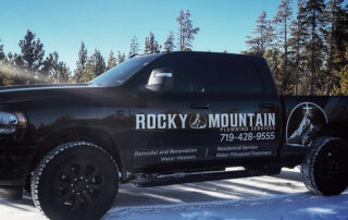 Black pickup truck with "Rocky Mountain Plumbing Services" logo and plumbing contact information, parked on snow with trees in the background.