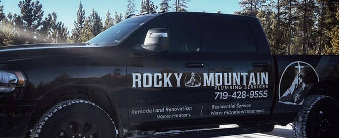 Black pickup truck with "Rocky Mountain Plumbing Services" logo and plumbing contact information, parked on snow with trees in the background.