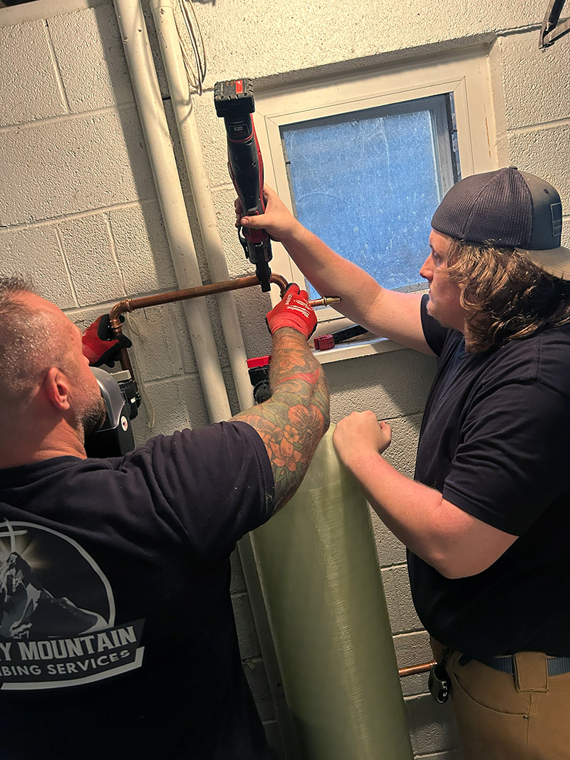 Rocky Mountain Plumbing Services Water Treatment System Install 800 Two plumbers work together on copper pipes near a window; one holds a pipe while the other uses a power tool to make plumbing adjustments.