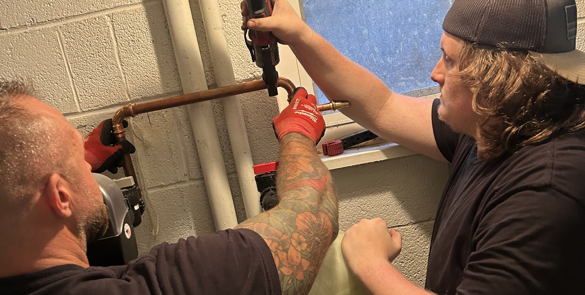 Two men use plumbing tools to work on a copper pipe near a window; one holds the pipe while the other tightens it with a pipe wrench.