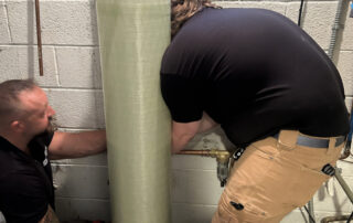 Two men work on plumbing behind a large, tall water filtration tank in a basement or utility room, ensuring the system runs smoothly.