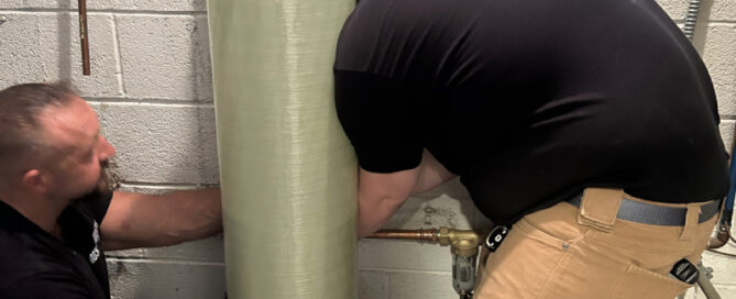 Two men work on plumbing behind a large, tall water filtration tank in a basement or utility room, ensuring the system runs smoothly.