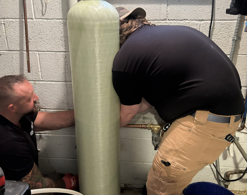 Water Softeners & Other Water Treatment Systems Two men work on plumbing behind a large, tall water filtration tank in a basement or utility room, ensuring the system runs smoothly.