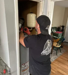 Why-Homeowners-Choose-Us A plumber wearing gloves and a "Rocky Mountain Plumbing" shirt works on water heater repair and installation in a small utility closet.